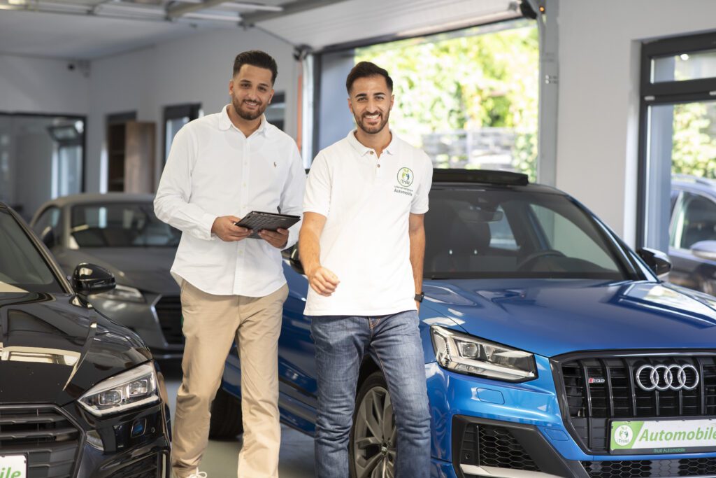 Zwei freundliche Mitarbeiter eines Gebrauchtwagenhändlers für Düsseldorf stehen in einem modernen Autohaus zwischen hochwertigen, geprüften Gebrauchtwagen verschiedener Marken – professionelle Beratung und Top-Service inklusive.